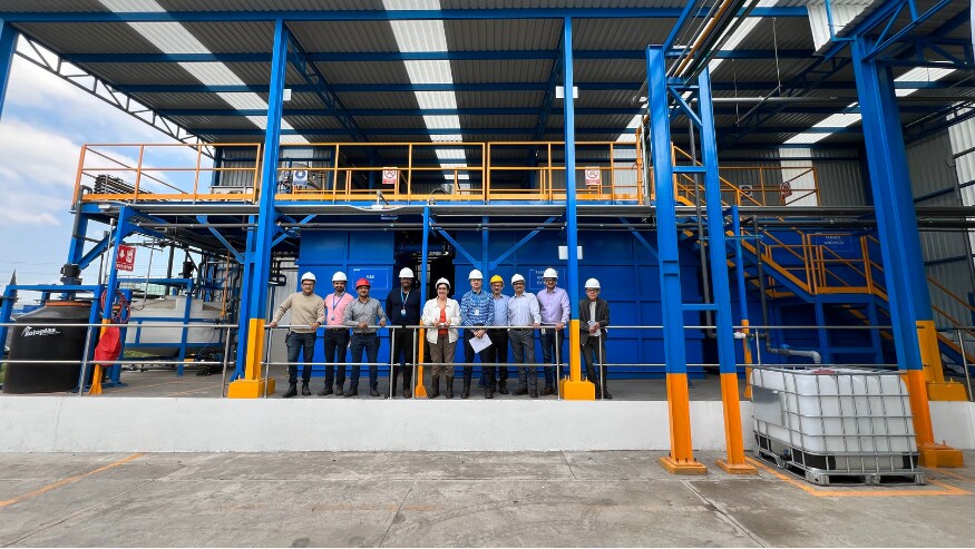 Nine people wearing safety helmets and reflective vests stand in front of an industrial facility with blue and orange beams