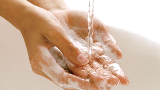 A person washing their hands and lathering soap over running water in a bathroom sink practicing hand hygiene.